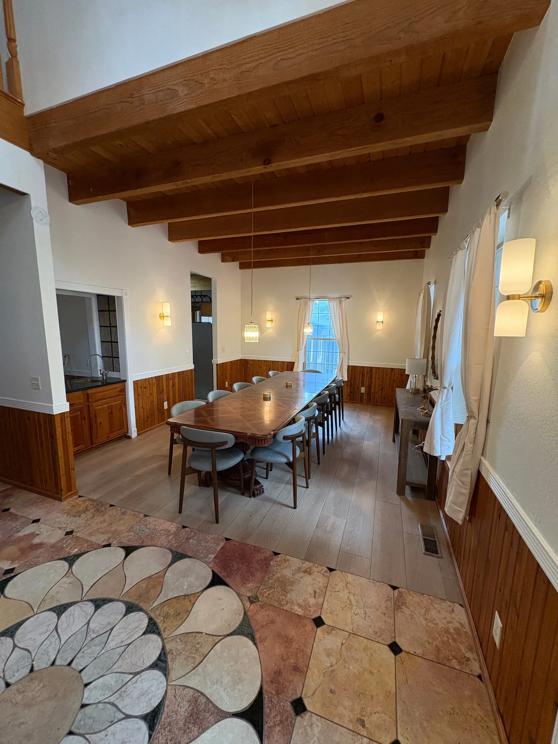 The dining room — one long polished table with ten upholstered chairs, under wall sconces and a beamed ceiling.