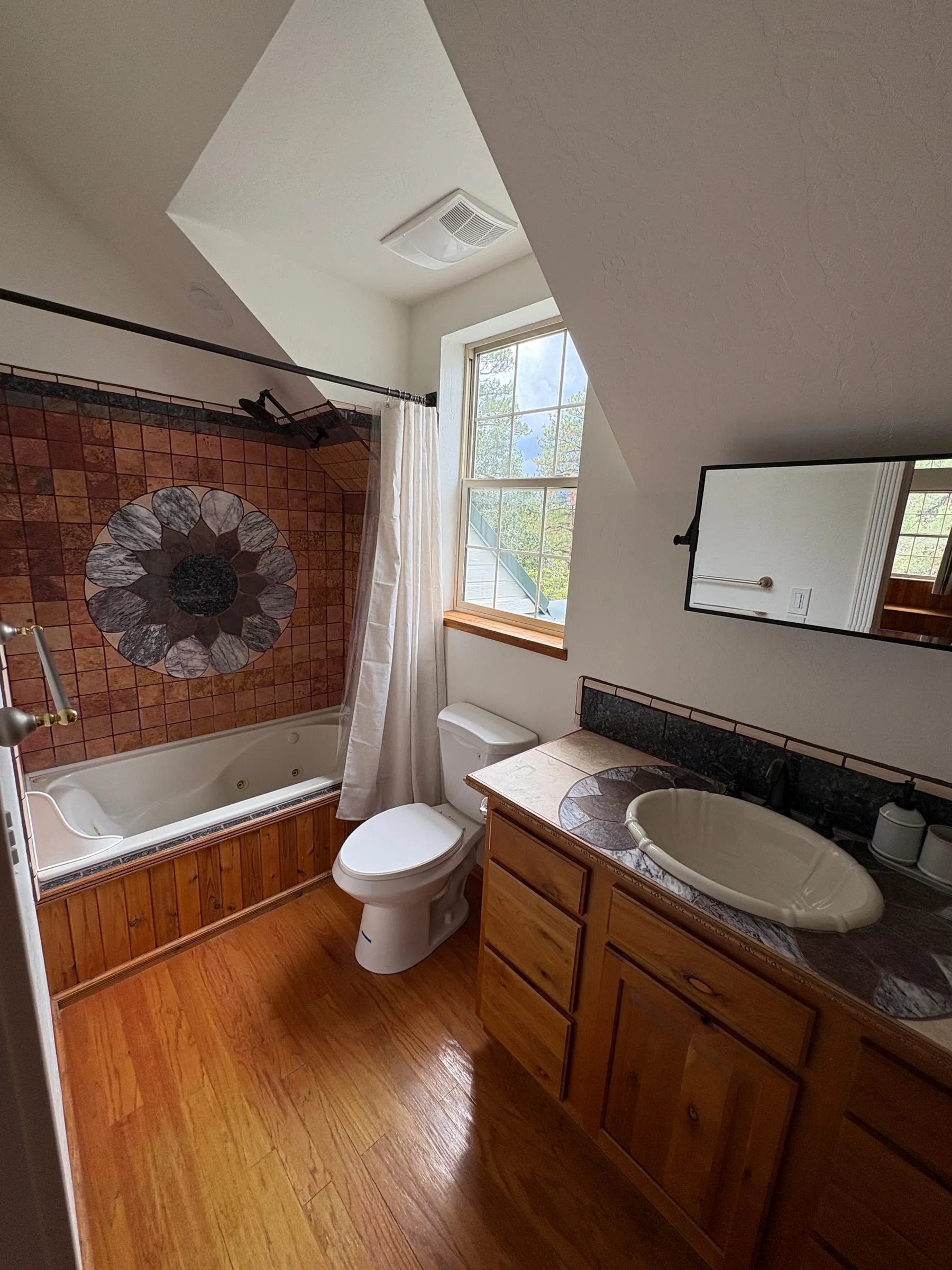 The ensuite bathroom in Harper’s Room — a corner tub set into tilework with a sunburst medallion, a wood vanity, and a window above the toilet.