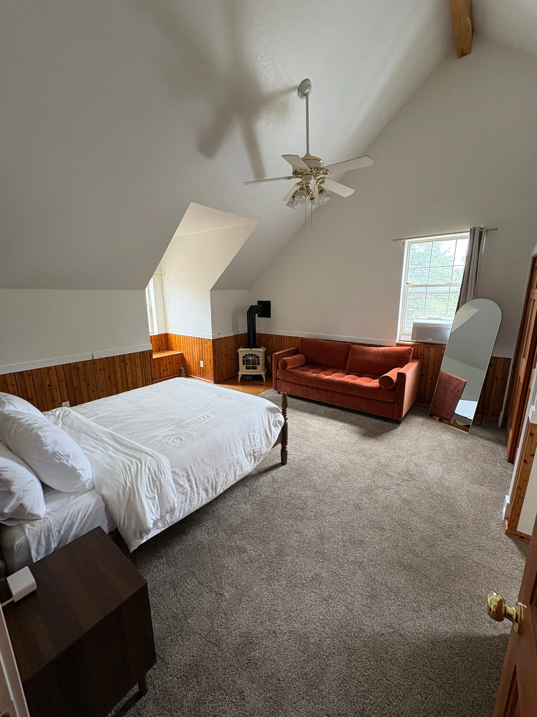 Harper’s Room — a queen bed and a blue sleeper sofa under a sloped ceiling, with a wood stove against the panelled wall and a built-in window seat at the far end.