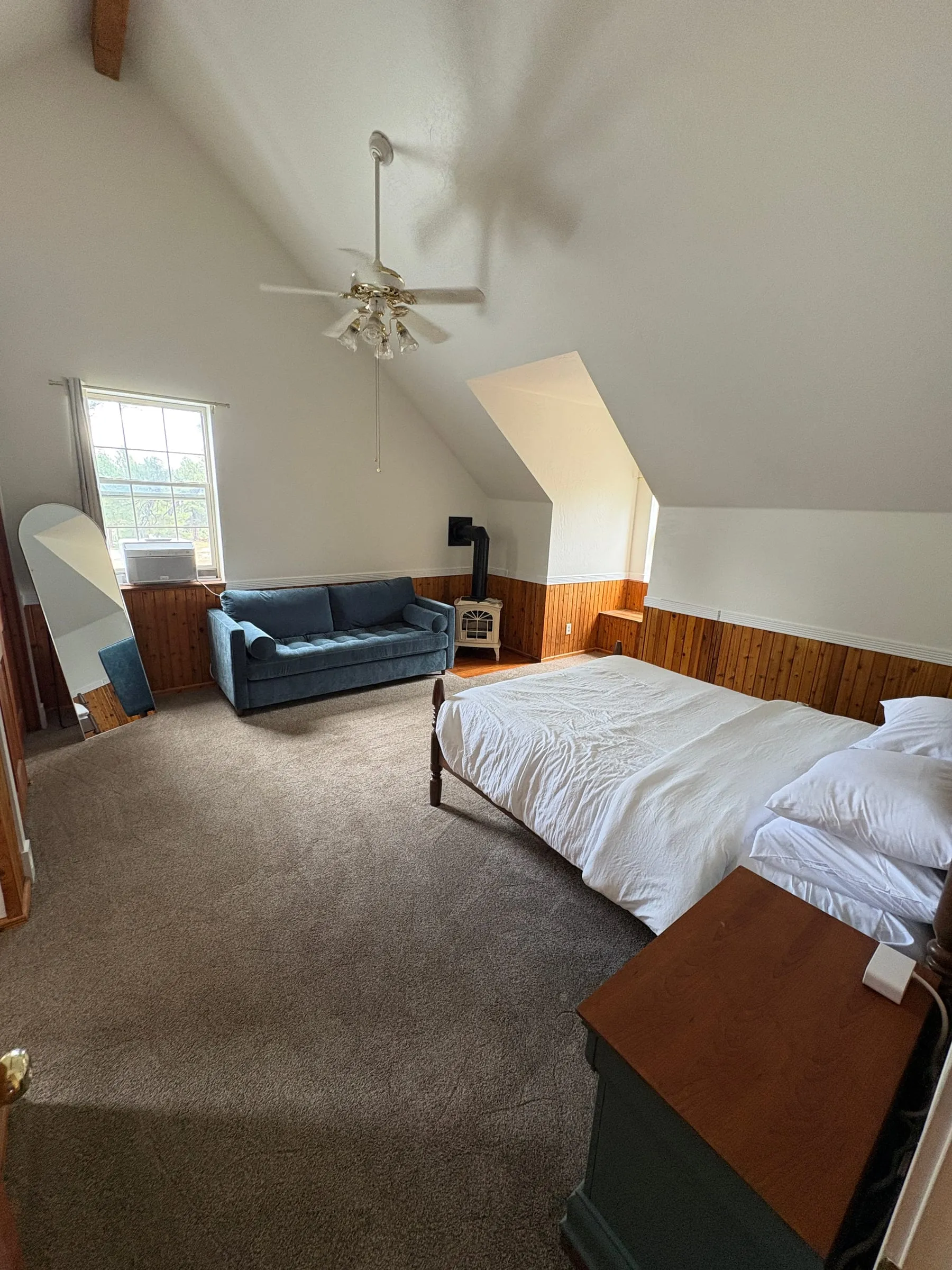 Jane’s Room — a queen bed and a blue sleeper sofa under a high sloped ceiling, with a cream-coloured gas stove against the panelled wall.