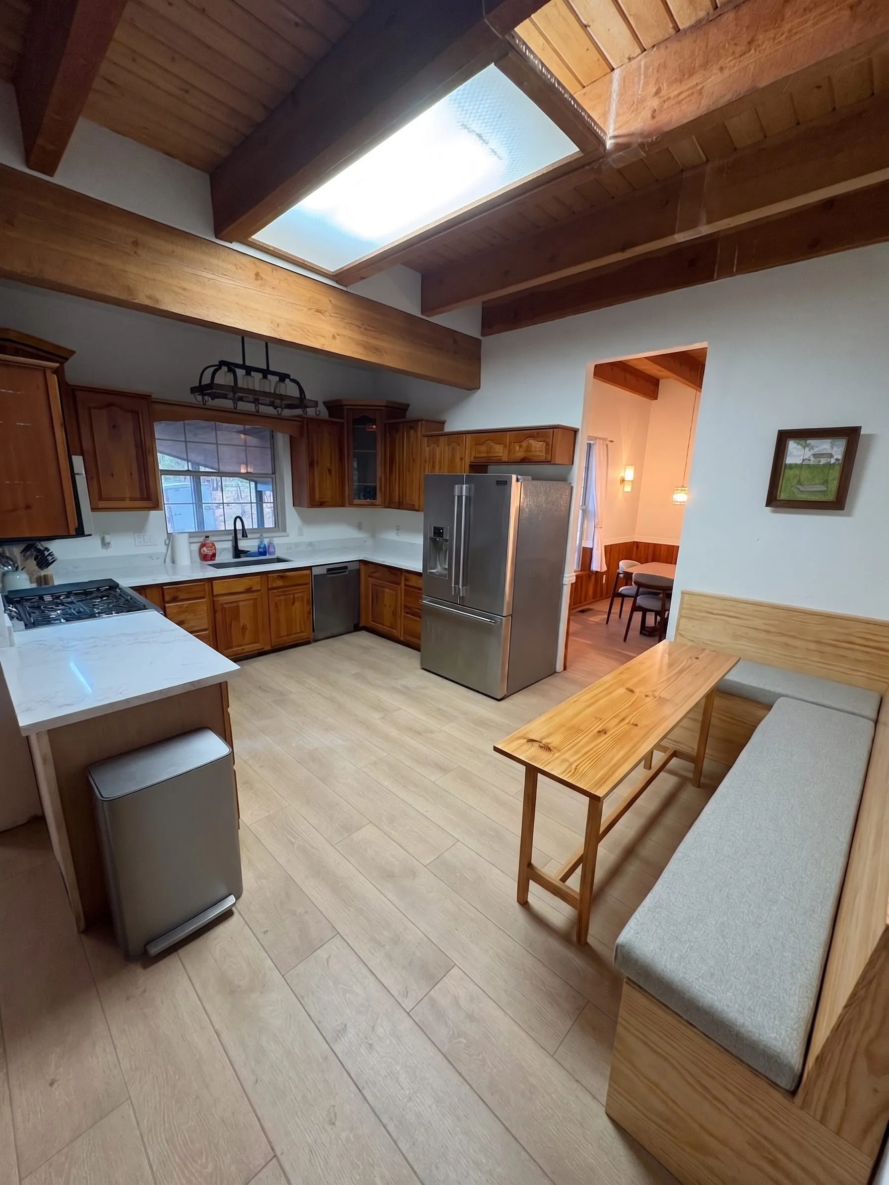 The kitchen — pine cabinets and a quartz island under a skylight and exposed beams, with a gas range, a stainless refrigerator, and a built-in banquette with a long wooden table.