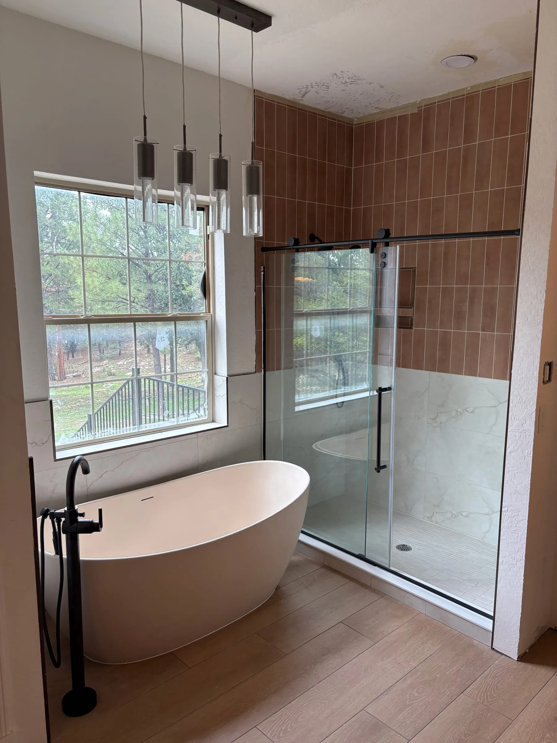 The ensuite bathroom in Madison’s Room — a white freestanding soaking tub with a floor-mounted filler, a large glass-walled shower with marble tile, and a tall window looking into the pines.