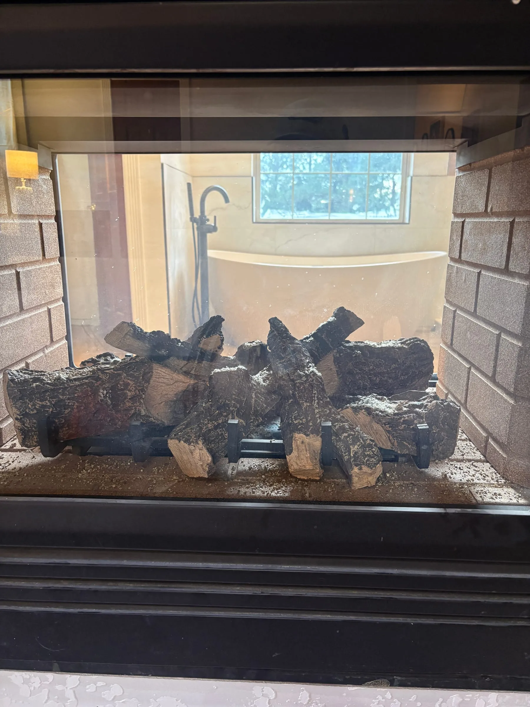 A gas fireplace in a carved oak mantel with a mirror above it. The firebox is open on both sides, so the bathroom is visible through the flames from the bedroom.