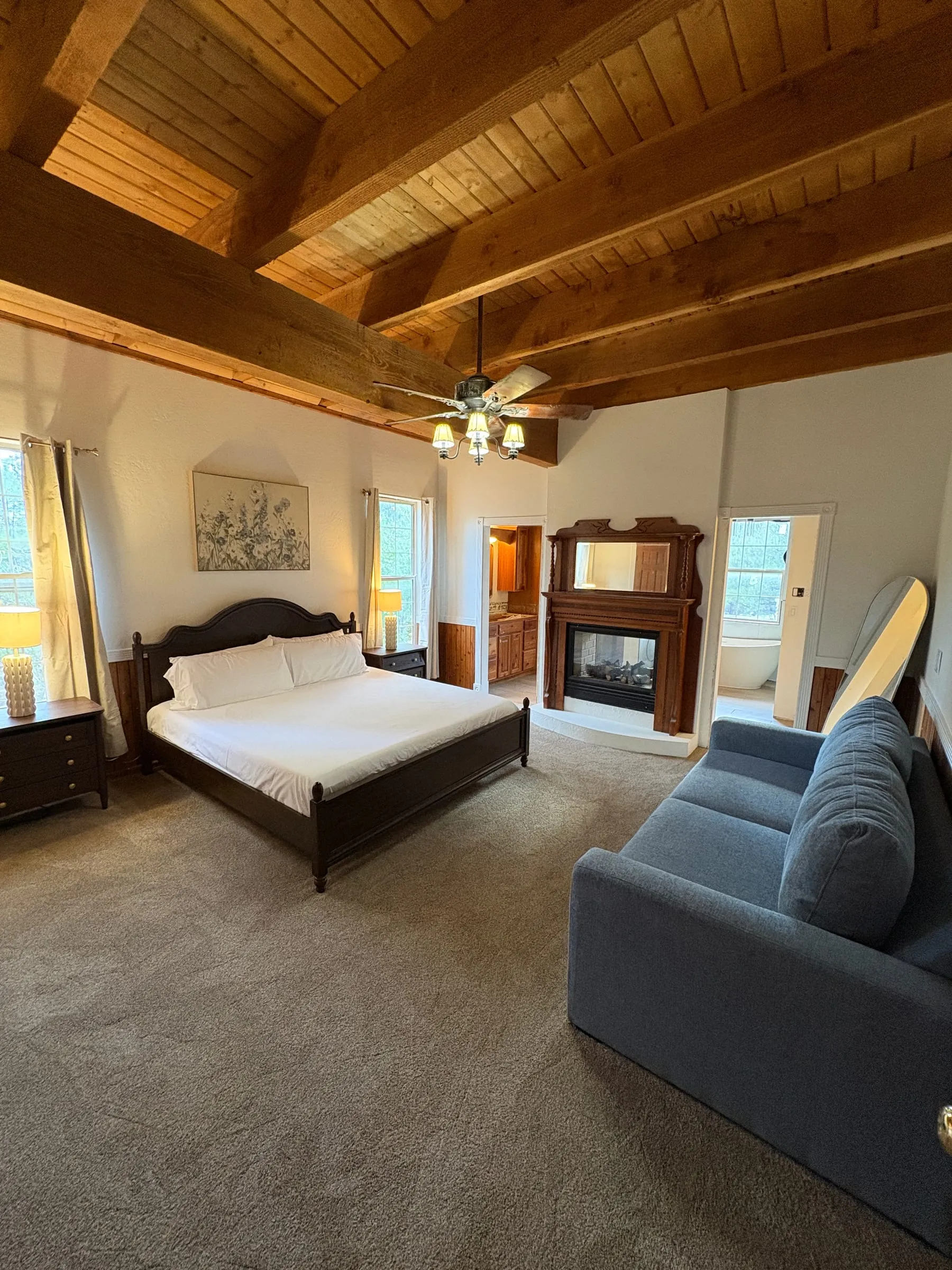Madison’s Room — a king bed with a dark carved headboard under a vaulted wood-beam ceiling. A blue sofa sits at the foot of the bed, and a fireplace is set into the far wall.