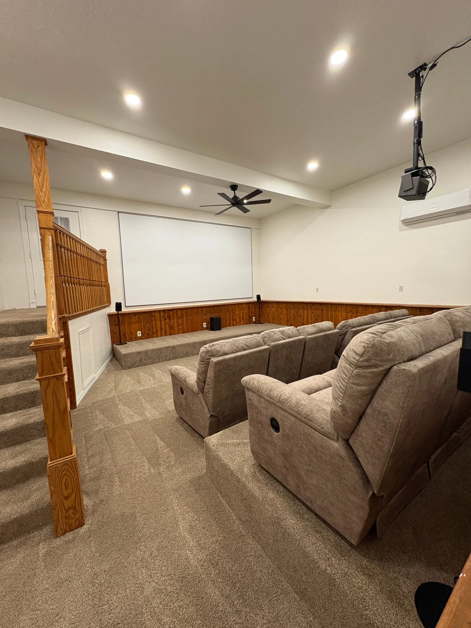 The theater — four full-size reclining couches facing a 200-inch screen, with a ceiling-mounted 4K projector, surround-sound speakers, and a small stage at the front.